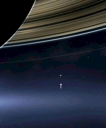 The image shows Saturn's rings with a small dot and an arrow pointing to it, possibly the Earth, signifying its tiny scale in space.