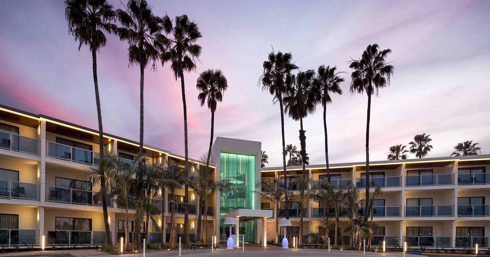 A mid-century hotel or motel with a glass central entrance, two floors of balconies, tall palm trees, and a colorful sunset sky.
