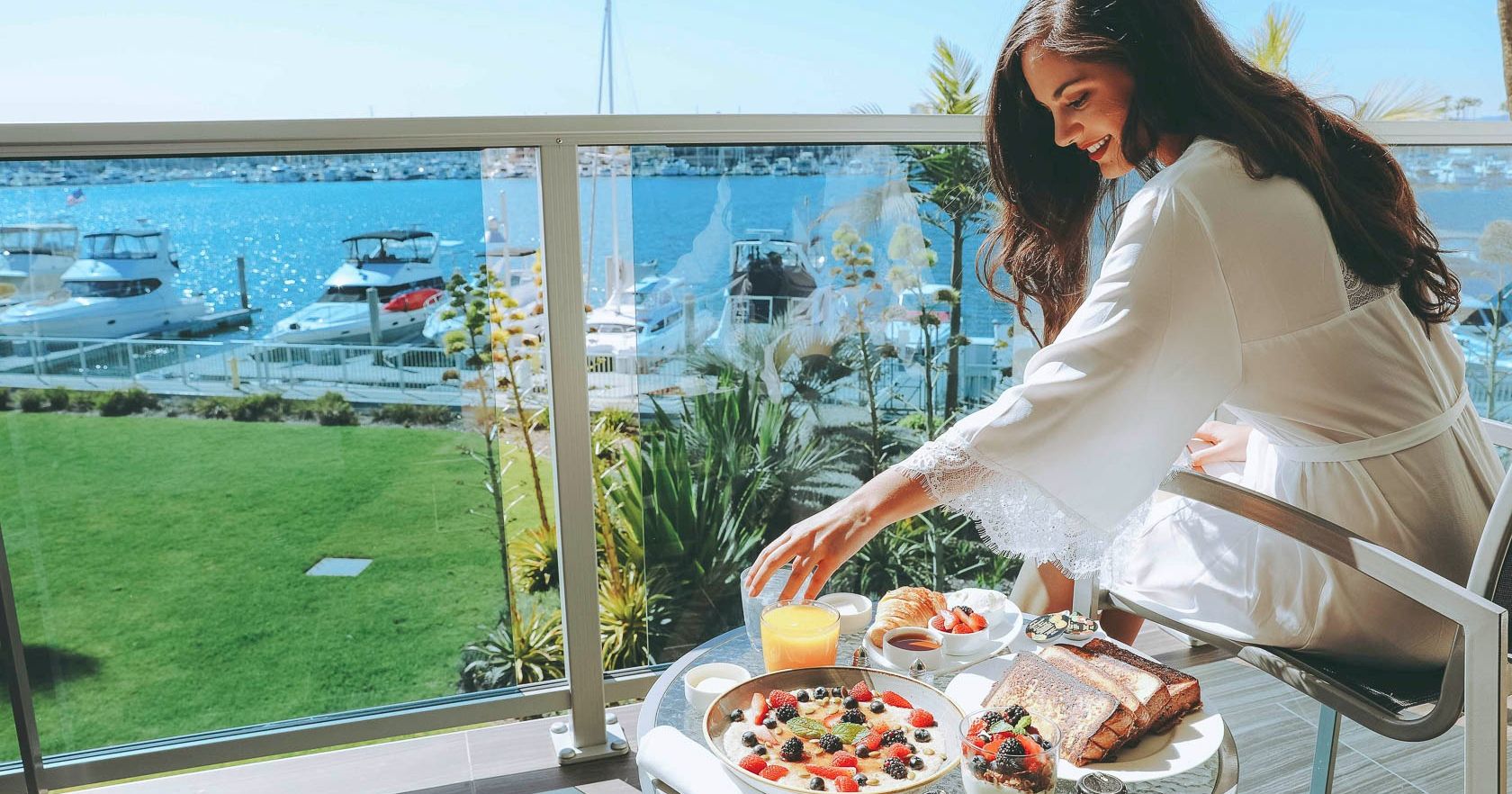 A woman enjoys a luxurious seaside breakfast on a balcony: cappuccino, pastries, berries, toasted bread, and a scenic marina view.