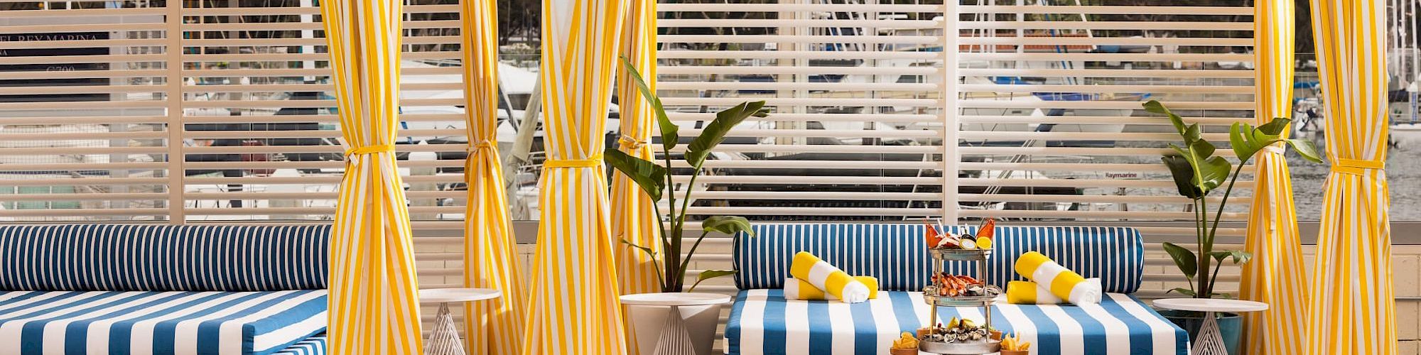 Two striped cabanas with yellow-and-white curtains by a pool, blue-and-white cushions, potted plants, and lounge chairs.