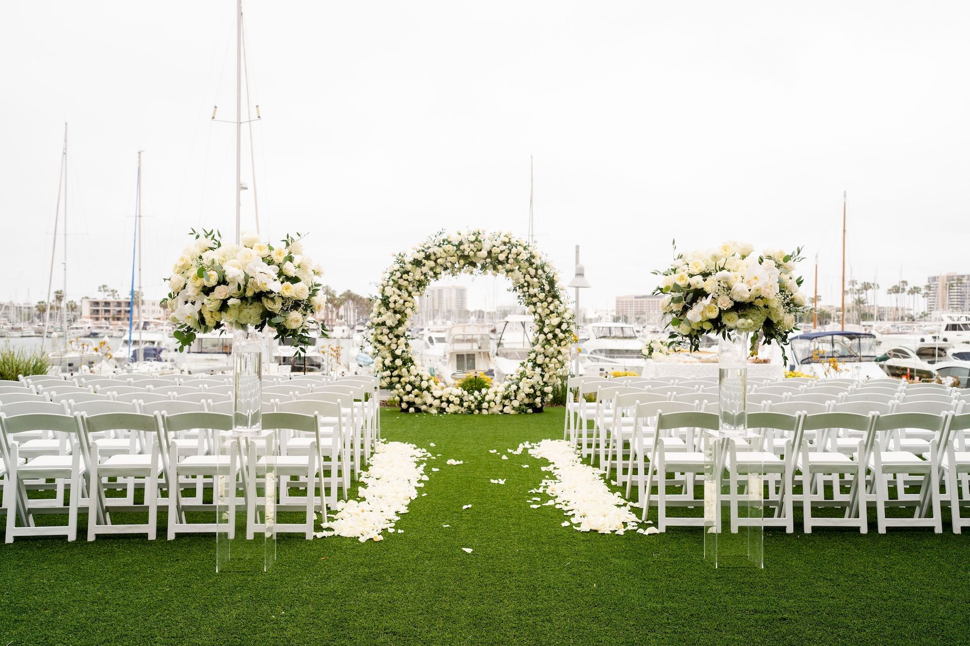A seaside wedding setup with white chairs, floral arch, and two tall flower arrangements lining a grassy aisle toward a circular floral arch, over water.
