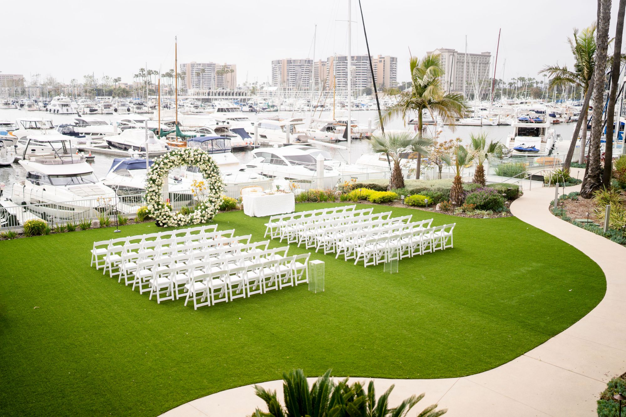 A grassy marina wedding setup with white chairs arranged in rows, a curved path, palm trees, and boats in the background.