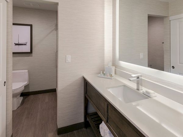 A modern bathroom with a double vanity, large backlit mirror, and a separate toilet area visible in the background, neutral tones throughout.