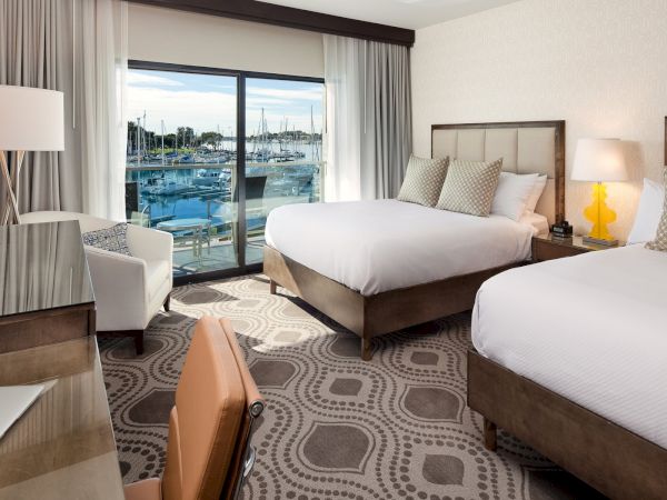 A modern hotel room with two queen beds, a desk and chair, a seating area by a window with a balcony, and soft lighting. The view shows water and boats outside.
