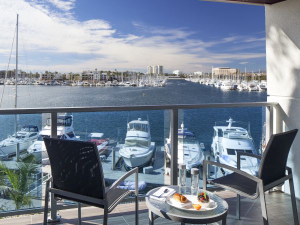 A balcony view over a marina with several boats, a glass table set with drinks and snacks, and two chairs facing the water.