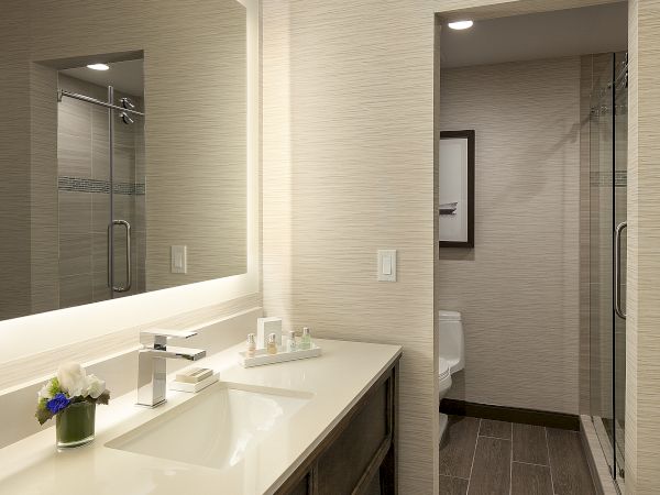 A modern bathroom with a long dual-sink vanity, large mirror, beige tiled walls, and a separate toilet/shower area visible in the background.