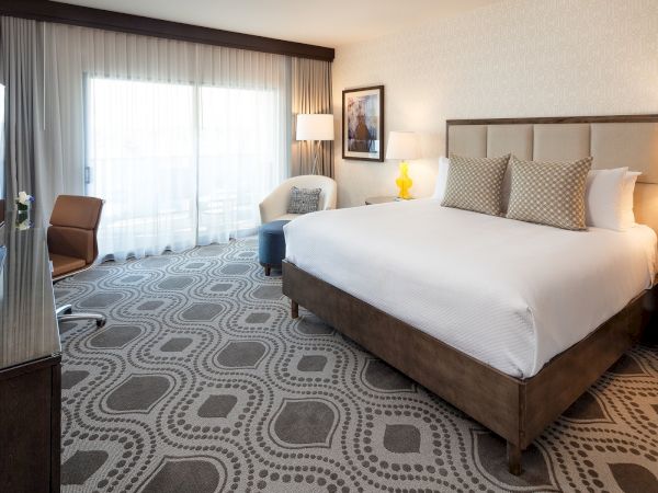 A modern hotel room with a king bed, two nightstands, lamps, a seating area by the window, and a patterned carpet.