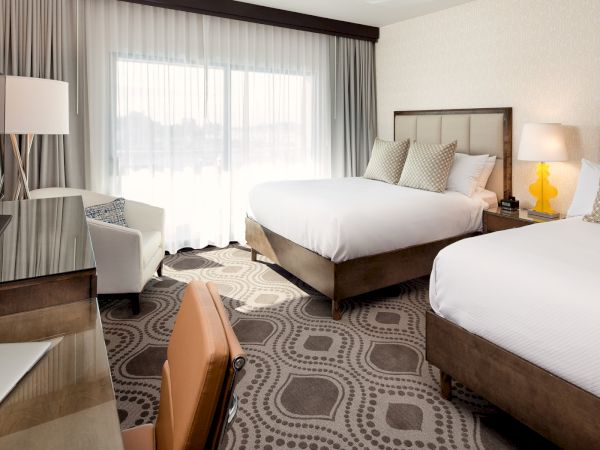 Two neatly made twin beds with brown wooden headboards, a desk with a laptop, a seating area, large windows with sheer curtains, and bedside lamps.