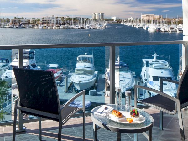 A sunny balcony view over a marina with several yachts, water, and distant buildings; a small table with drinks and snacks, two chairs.