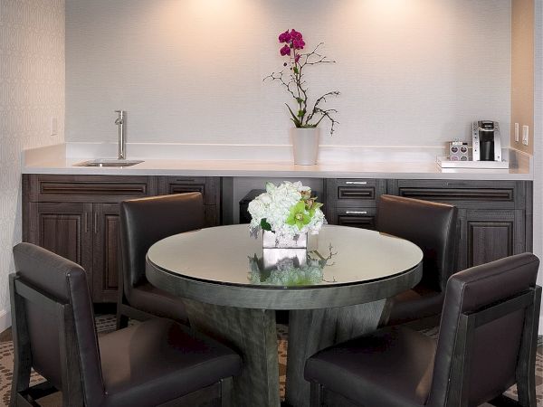 A modern kitchenette with a round glass dining table, six black chairs, a sleek gray counter, and a single flower vase centerpiece.