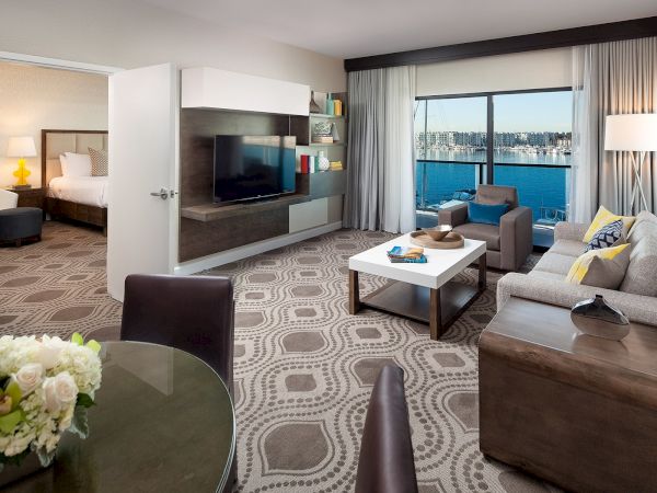 Spacious hotel suite with a living area, large windows, city or water view, modern furnishings, a dining table, and an adjacent bedroom.