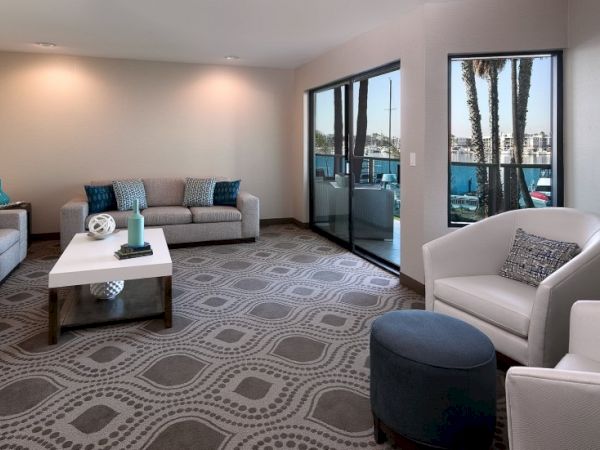 A bright living room with a gray patterned rug, beige sofas, white chairs, a white coffee table, and large glass doors opening to a balcony.