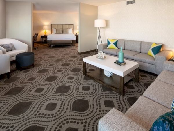 A modern hotel suite: a seating area with sofa, chairs, coffee table, decorative pillows, and a view into a bedroom with a bed and lamps.