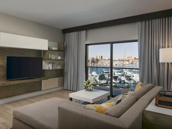 A modern living room with a wall-mounted TV, wooden console, built-in shelves, a gray sectional sofa, a coffee table, and a large window overlooking a marina with boats.
