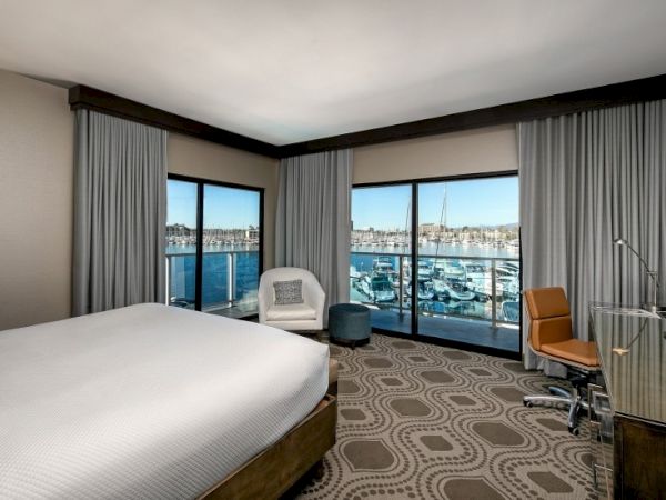 A modern hotel room with a large bed, a seating area, and floor-to-ceiling windows overlooking a marina, desk, and neutral d&eacute;cor.