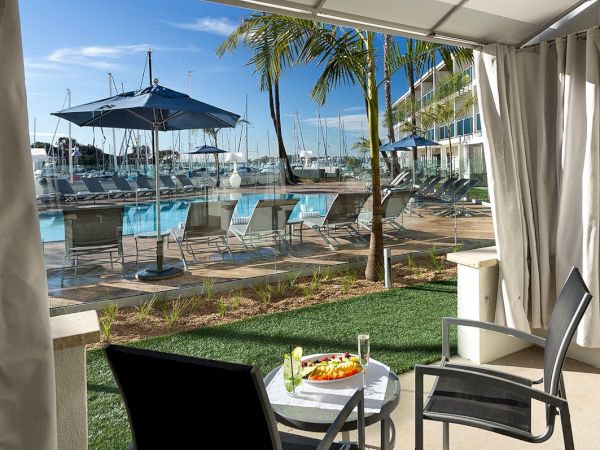 A balcony view over a marina with blue lounge chairs, palm trees, a sun umbrella, and a table set with a plate of fruit, sea beyond.