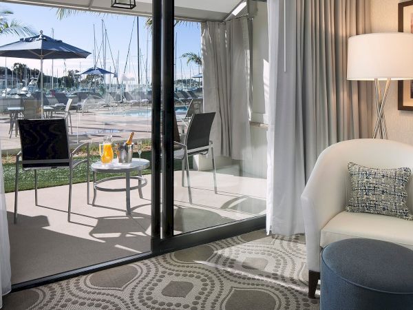 A luxurious hotel suite with a seating area opening to a balcony overlooking a marina, glass doors, and a patterned rug.