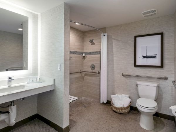 A clean, accessible hotel bathroom with a long sink, mirror-lit vanity, walk-in shower, toilet, and a framed nautical print on the wall.