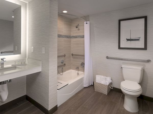 A modern hotel bathroom with a single sink, large mirror, a bathtub with a shower curtain, a toilet, a towel basket, and a framed picture on the wall.