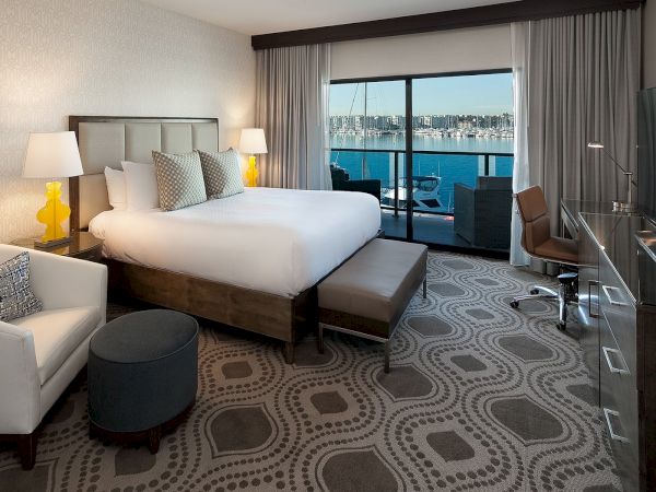 A modern hotel room with a large bed, two nightstands, a sofa, ottoman, and a desk; sliding doors open to a balcony with a waterfront view.