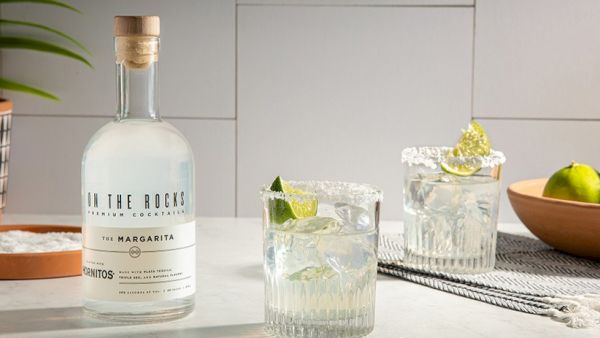 A bottle of tequila in the foreground with two on-the-rocks cocktails, a lime garnish, a wooden bowl of limes, and a potted plant in a modern kitchen backdrop.