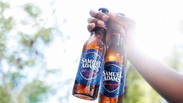 Two people clinking Samuel Adams beer bottles outdoors, raised in a toast against a bright, sunny background. End the sentence.