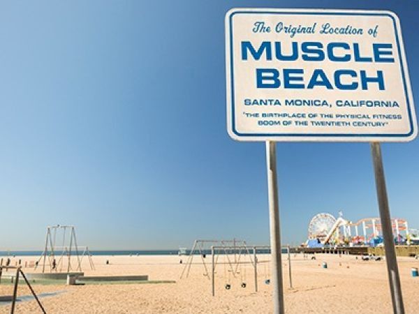 Muscle Beach sign on a sunny Santa Monica beach, with swings, sand, and a distant Ferris wheel by the boardwalk.