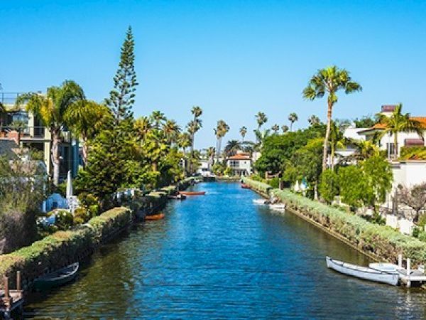 A sunny canal lined with palm trees and houses on both sides, calm blue water, small boats docked along the edges, clear sky.