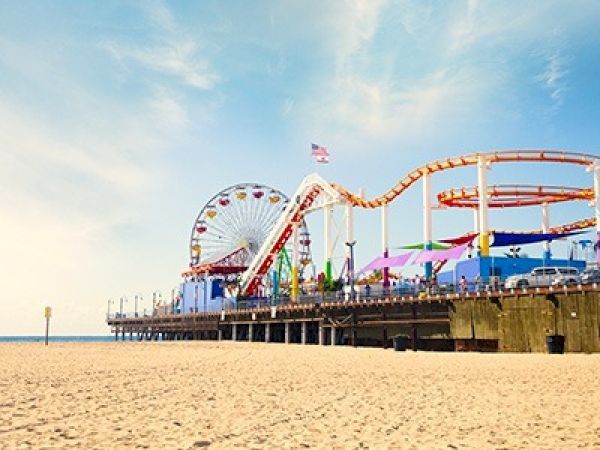 A sunny beach with a wooden pier and a colorful amusement park roller coaster and ferris wheel extending over the sand and water.