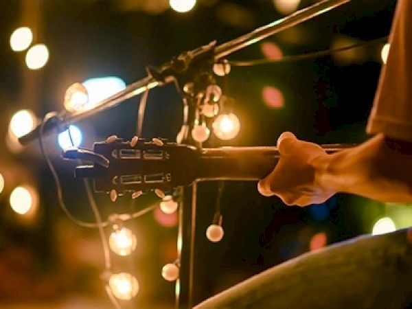 A musician&rsquo;s hand on a microphone stand with guitar cables and warm string lights in the background at a dim stage, creating a cozy concert vibe.