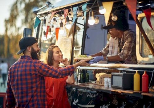 A couple buys food from a vendor at a colorful food truck, the vendor handing them a dish while festive lights and bunting glow behind them.