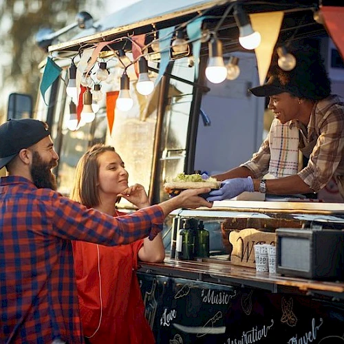 A couple buys food from a vendor at a colorful food truck, the vendor handing them a dish while festive lights and bunting glow behind them.