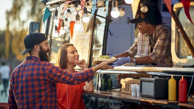 A couple buys food from a vendor at a colorful food truck, the vendor handing them a dish while festive lights and bunting glow behind them.
