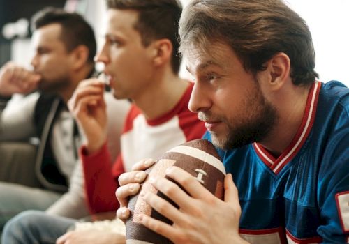 Three men watching a game, one holding a football and focused, others in the background sipping drinks, all seated.