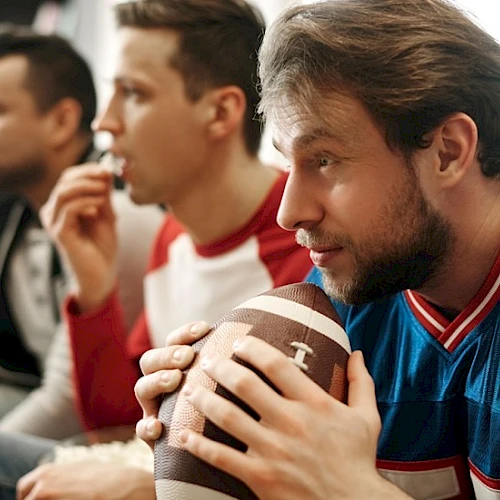 Three men watching a game, one holding a football and focused, others in the background sipping drinks, all seated.