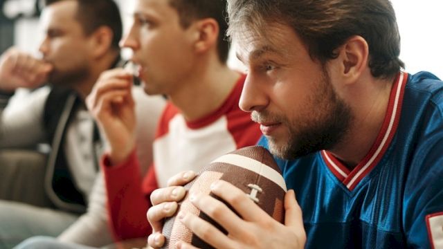 Three men watching a game, one holding a football and focused, others in the background sipping drinks, all seated.