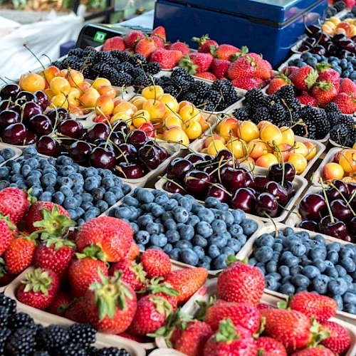 Colorful berries and stone fruits arranged in neat crates at a market, with ripe strawberries, blueberries, cherries, and apricots. Top it at 140 characters, always ending the sentence.