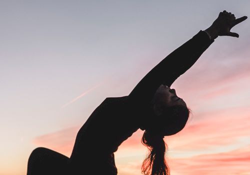A person doing a backbend against a sunset sky, arms stretched overhead, silhouette in a graceful athletic pose at dusk.