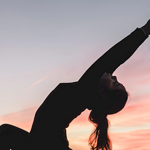 A person doing a backbend against a sunset sky, arms stretched overhead, silhouette in a graceful athletic pose at dusk.