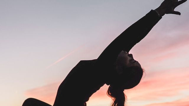 A person doing a backbend against a sunset sky, arms stretched overhead, silhouette in a graceful athletic pose at dusk.