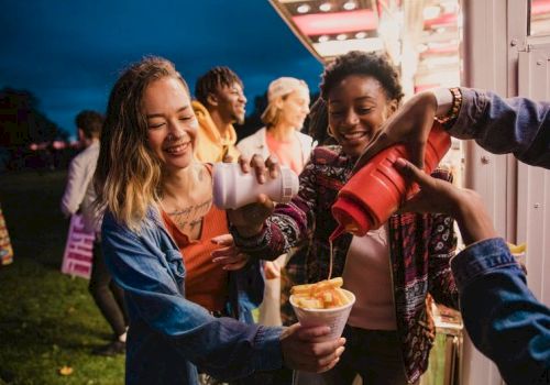 Friends at a carnival booth share smiles as drinks and snacks flow, laughter all around while someone fills cups with treat-filled ice cream.