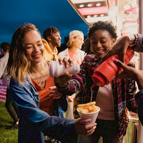 Friends at a carnival booth share smiles as drinks and snacks flow, laughter all around while someone fills cups with treat-filled ice cream.