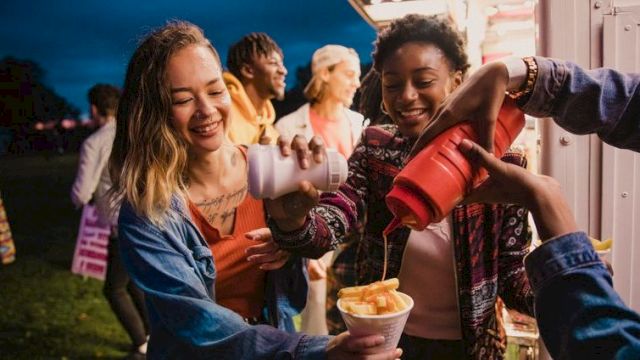Friends at a carnival booth share smiles as drinks and snacks flow, laughter all around while someone fills cups with treat-filled ice cream.