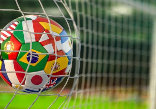 A soccer ball with multiple national flags printed on it is stuck in a goal net, close-up, on a grassy field.
