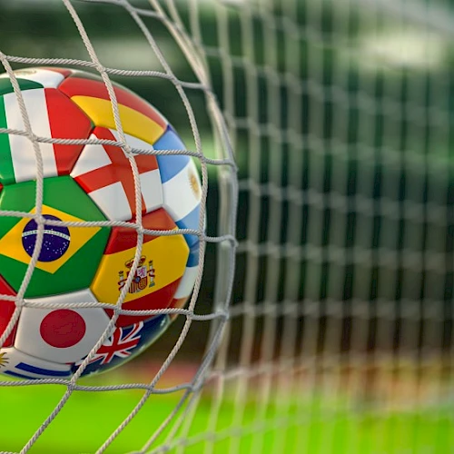 A soccer ball with multiple national flags printed on it is stuck in a goal net, close-up, on a grassy field.