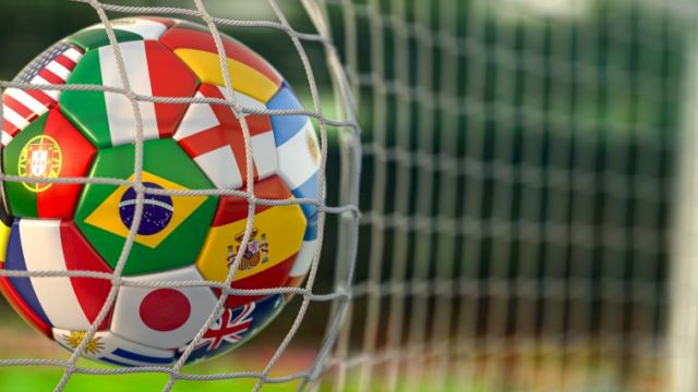 A soccer ball with multiple national flags printed on it is stuck in a goal net, close-up, on a grassy field.