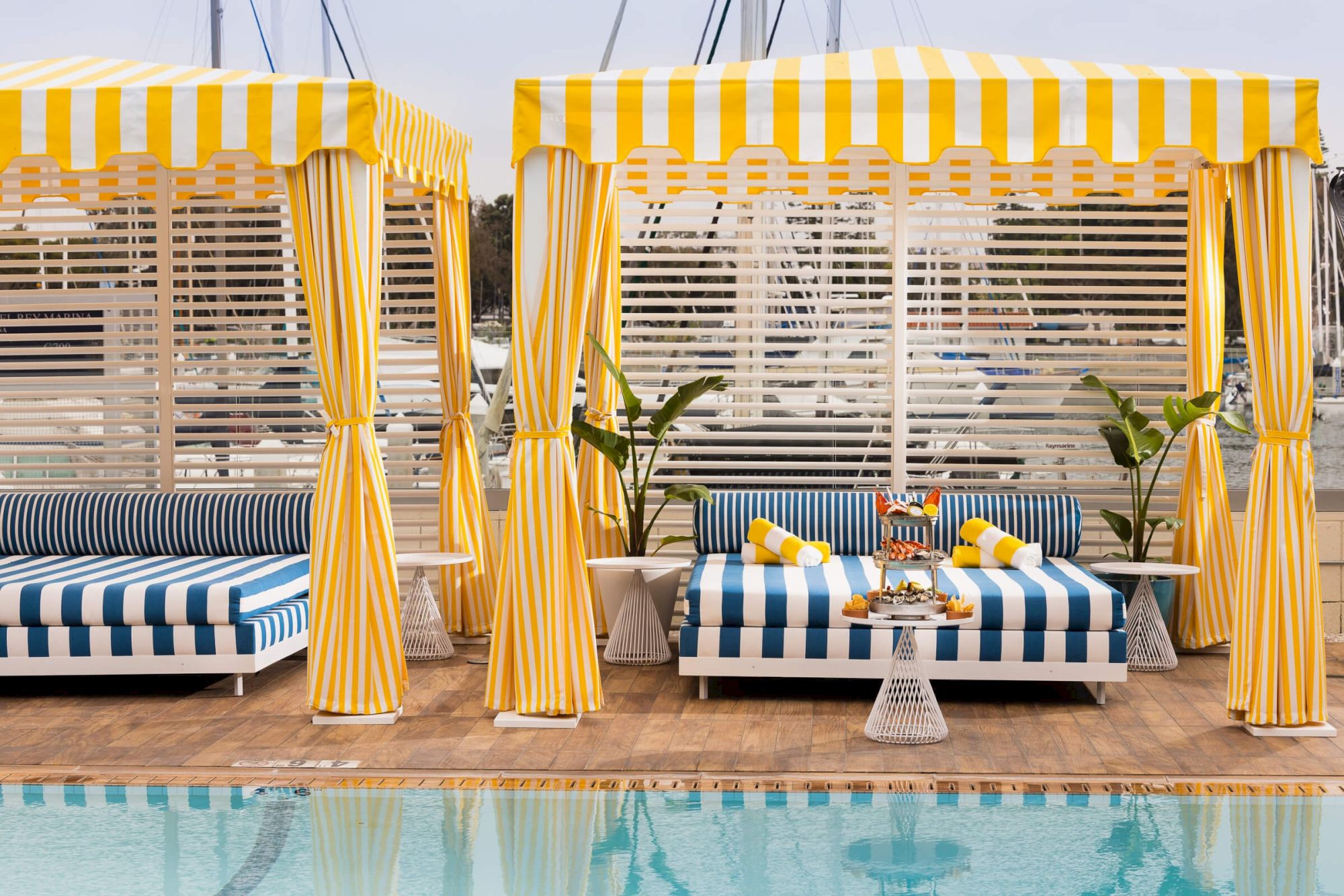 A resort-style pool area with yellow striped cabanas, blue-and-white loungers, potted plants, and a bright, sunny vibe.