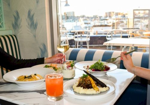 Two people dine at a bright caf&eacute; by the window, sharing drinks and plates of pasta and salad on a white-table setting.