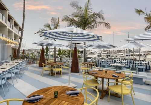 A sunny beachside cafe with wooden tables, yellow chairs, blue-striped umbrellas, and palm trees, ready for diners at sunset.