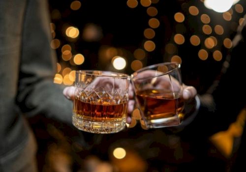 Two people clink whiskey glasses in a dim, festive setting with warm bokeh lights in the background.